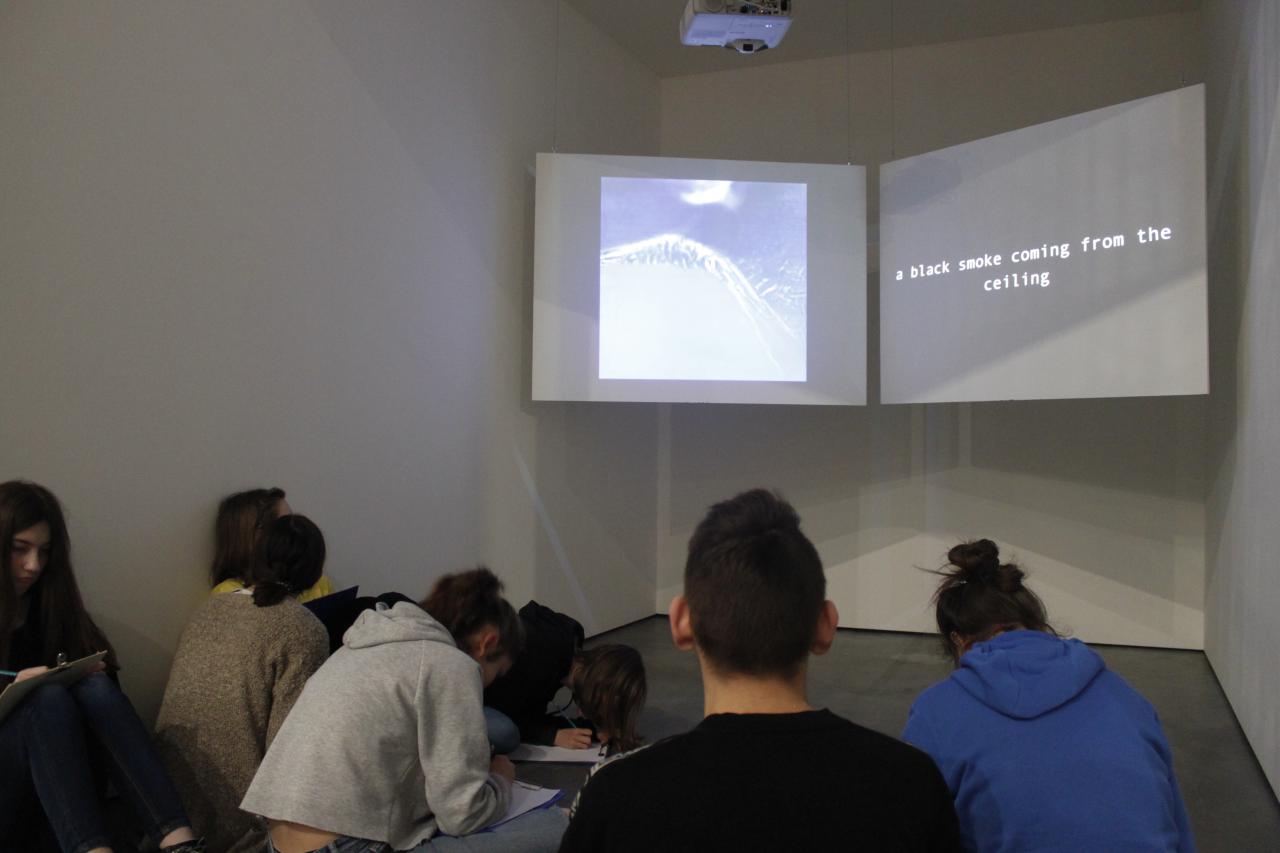Young people sit in front of two large projection screens on which a picture and the line "a black smoke coming from the ceiling" can be seen.