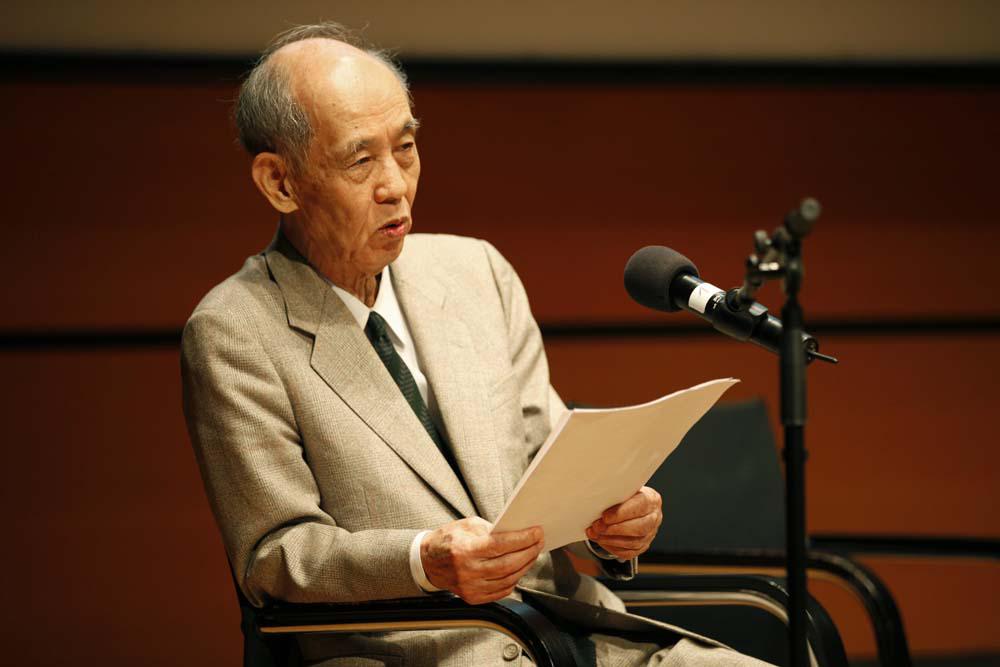 Hiroshi Kawano during his presentation. He is wearing a light suit, in his hands he holds his manuscript.