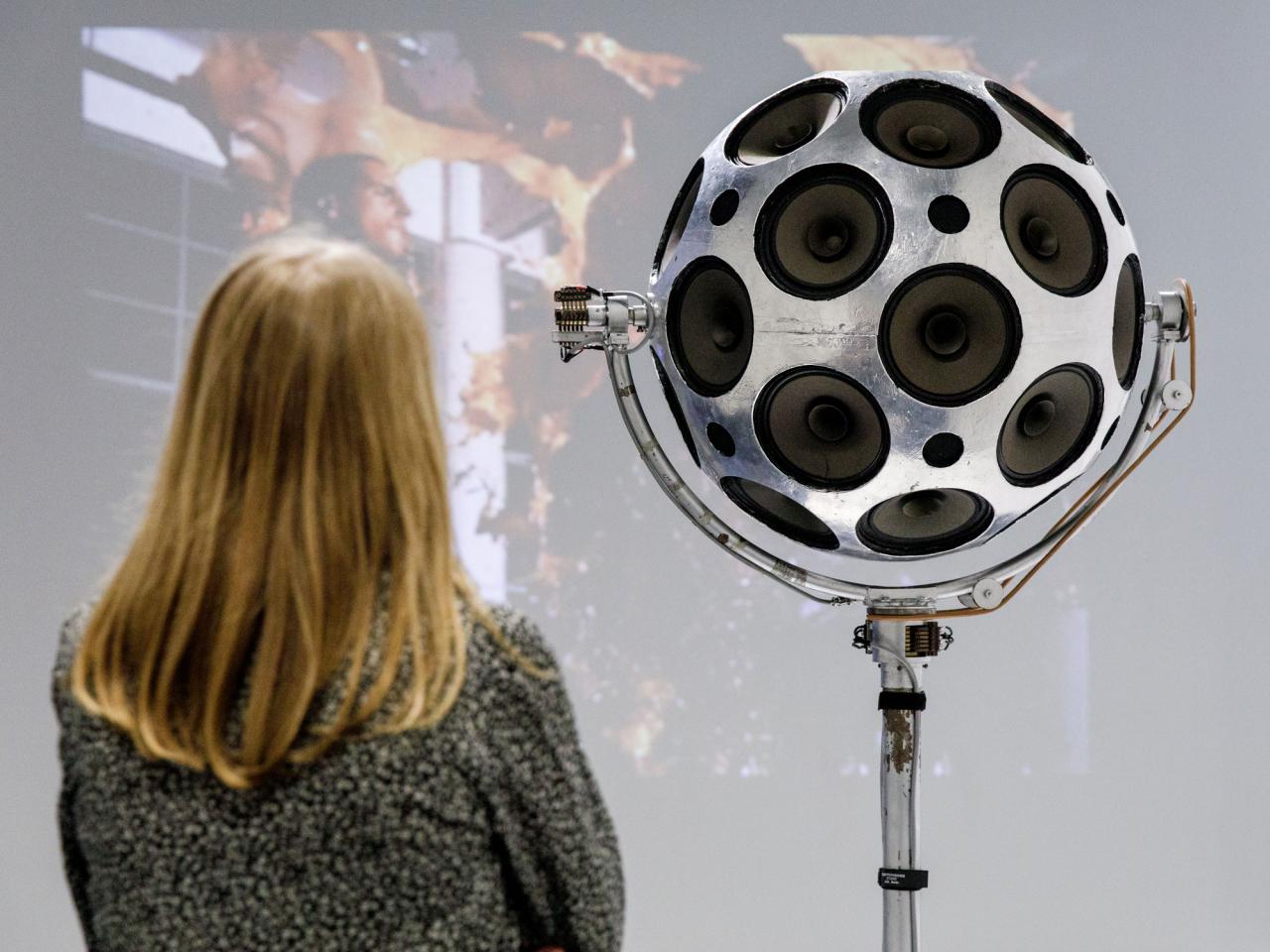 A woman stands in front of a silver sphere of loudspeakers