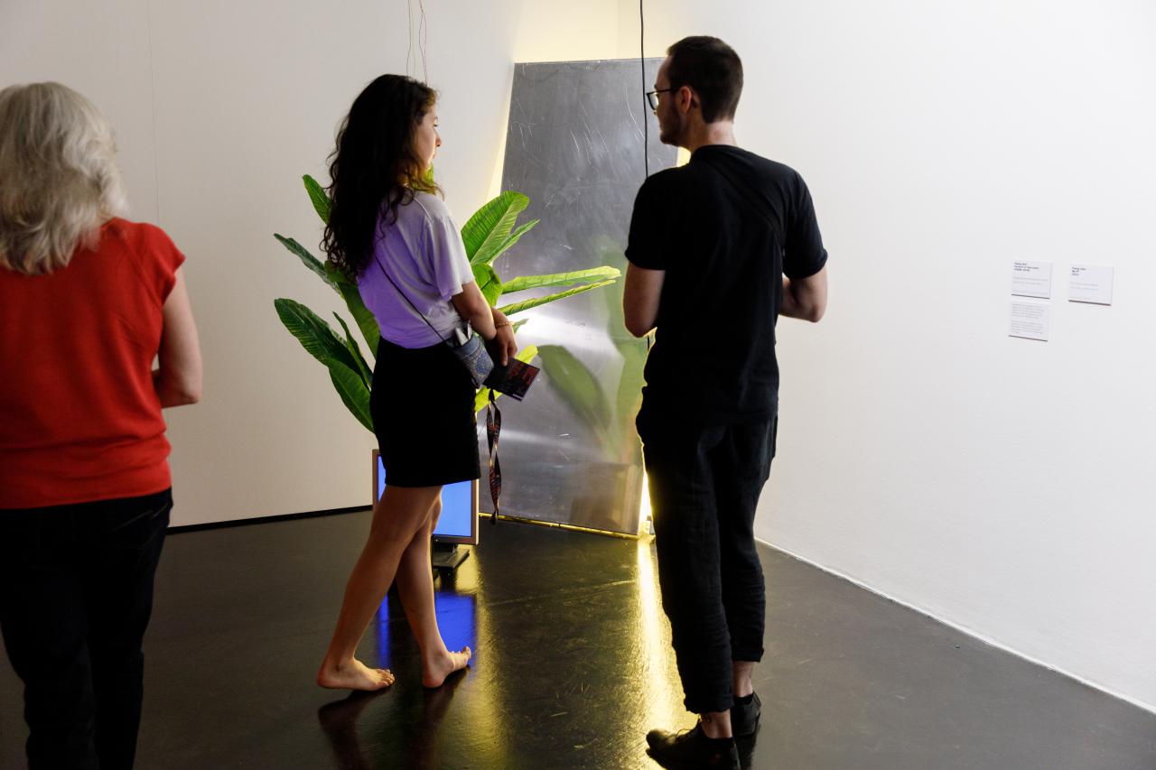 Das Foto zeigt eine barfüßige Besucherin und einen schwarz gekleideten Besucher vor einer an der Wand angelehnten Installation neben der eine Pflanze steht
