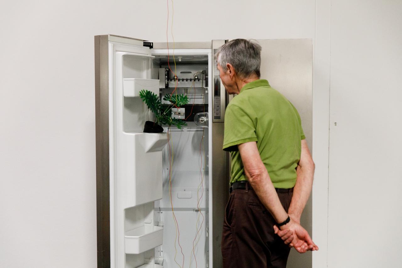 An elderly gentleman stands in front of an open refrigerator, which has been converted into a media installation. 