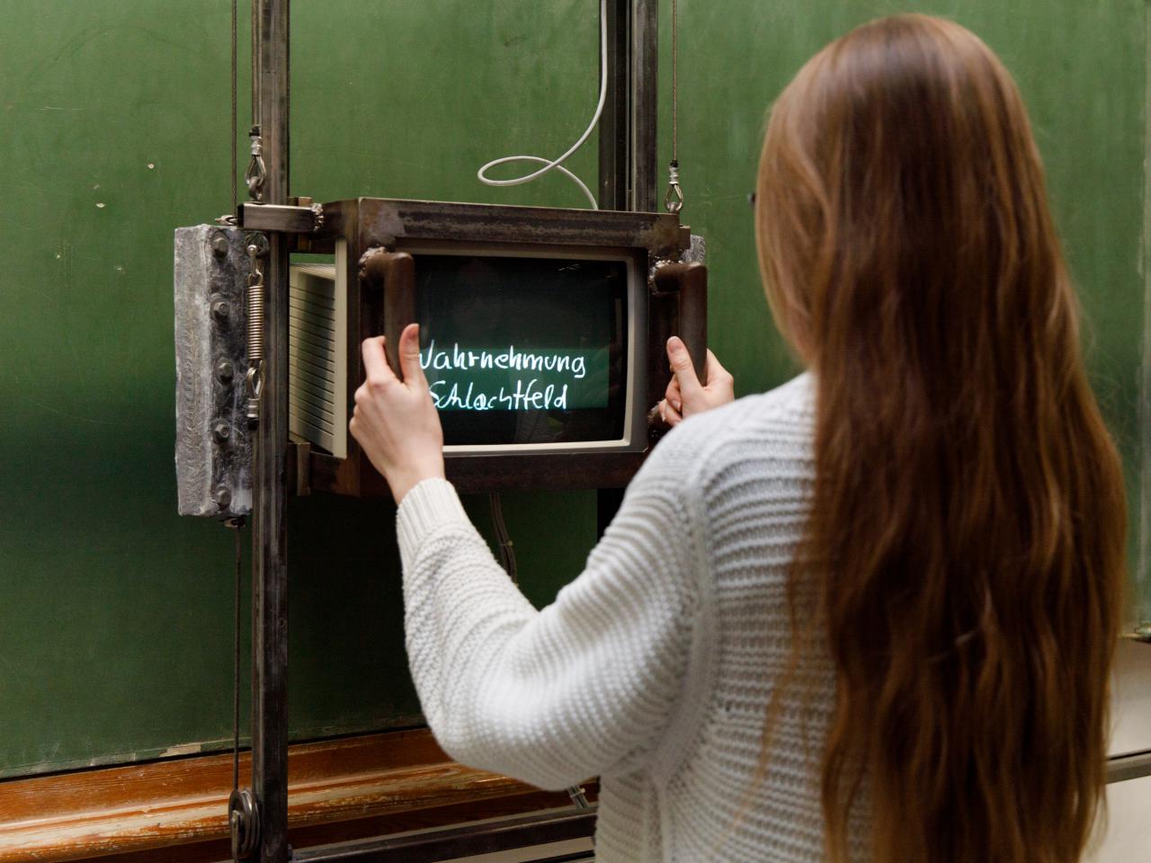 Besucherin vor Fietzeks »Tafel«, eine leere grüne Schultafel, auf der mit Hilfe eines Monitors zuvor unsichtbare Texte auftauchen.