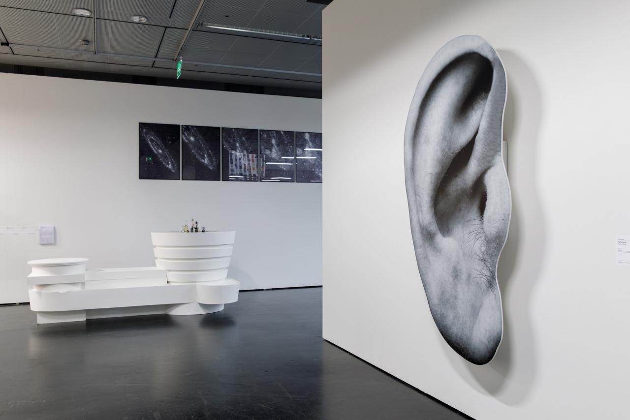 On the left, a white installation that looks like a futuristic bar. In the background black and white images of the universe. On the right half of the picture a wall with an oversized pressure of an ear.