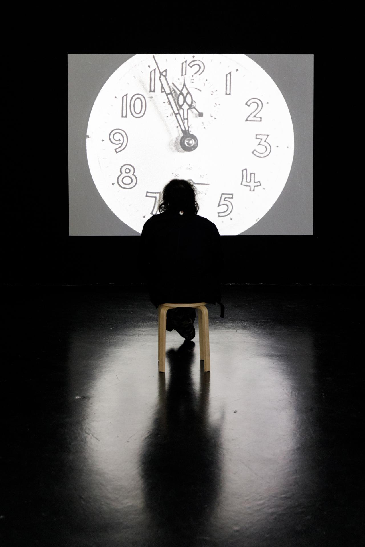 The photo shows the silhouette of a person watching a black and white film on a large screen. On the screen, an analogue clock strikes twelve. The light of the screen falls a round blurred light on the floor.