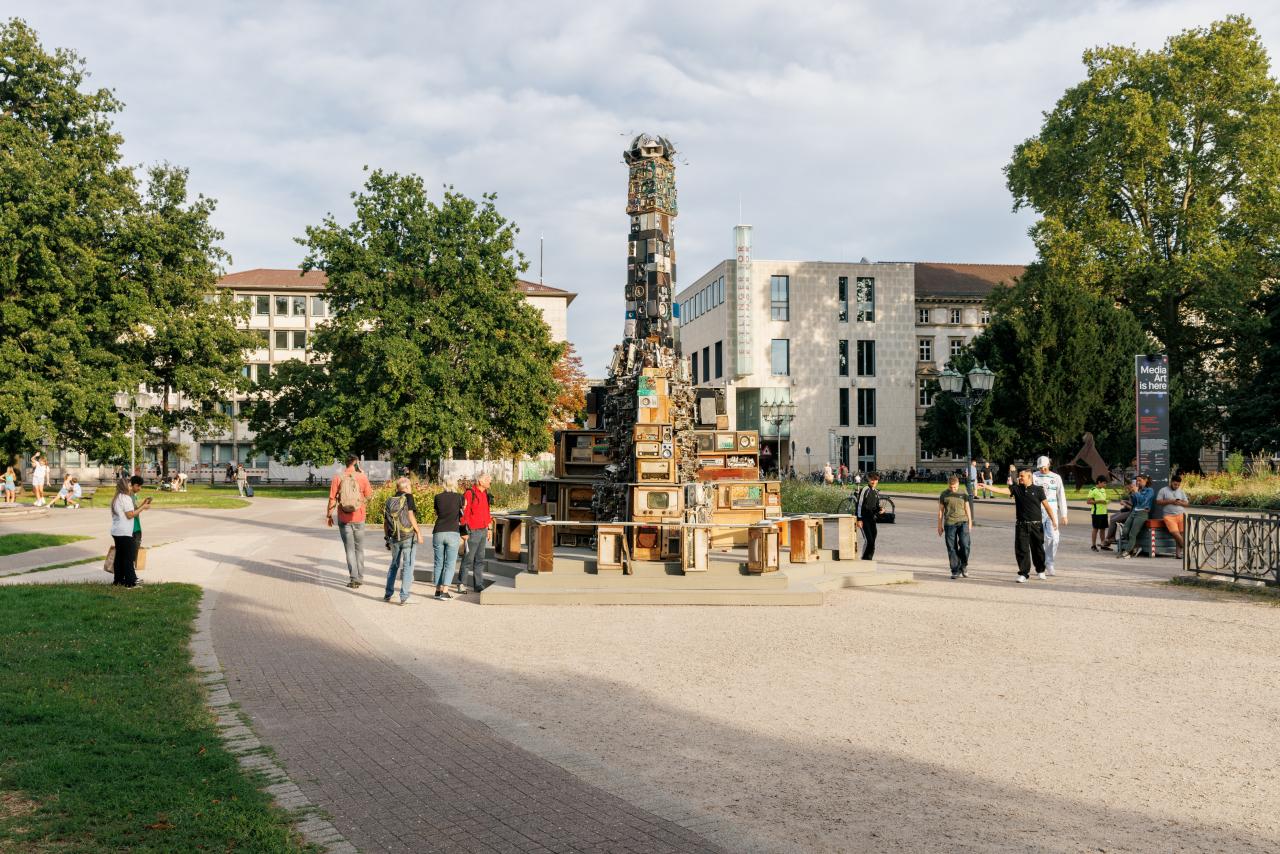 Auf dem Bild zu sehen ist die Medieninstallation »STREAMERS – a COVID Sculpture« von Benoît Maubrey auf Friedrichsplatz. Ein großer Turm von alten Lautsprecher ragt in die Höhe und viele Menschen haben sich darum versammelt um es sich anzusehen.