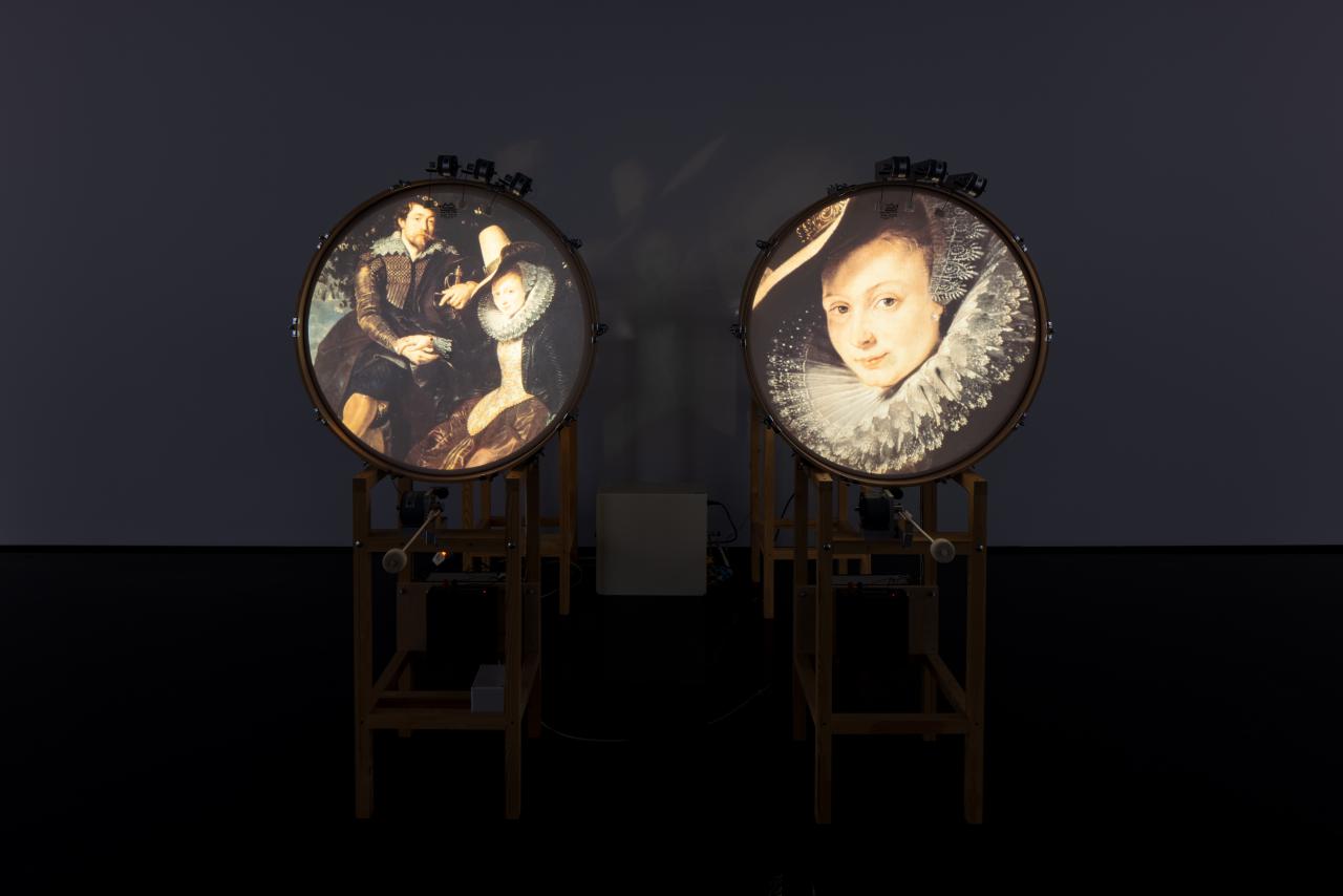 Zu sehen sind zwei stehende Trommeln, welche jeweils ein Bild auf der runden Fläche zeigen. Links ein altertümliches Ehepaar und rechts das Porträt einer Frau. 