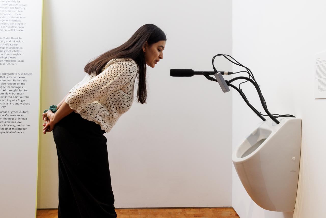 Zu sehen ist eine Person, die in ein Mikrofon spricht. Das Mikrofon ist an einer Toilette befestigt.