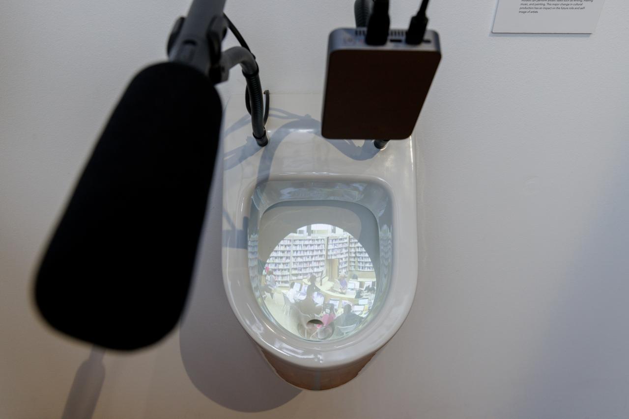 You can see a toilet bowl photographed from above. In it there is a projection. A microphone is fixed on the toilet.