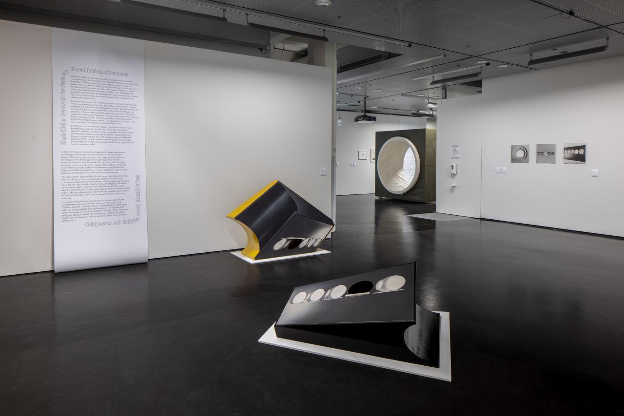 In the exhibition space, two sculptural works can be seen in the foreground. In the background, text is written on the wall and other works can be seen. 