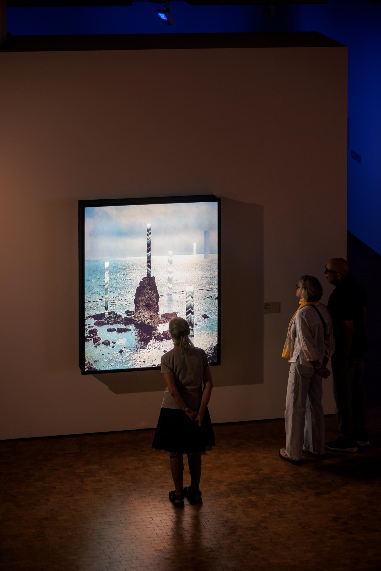 Zu sehen sind drei Menschen, die sich ein Kunstwerk anschauen, welches an der Wand hängt. Auf dem Kunstwerk zu sehen ist etwas, das an einen Eisberg in der Arktis erinnert.