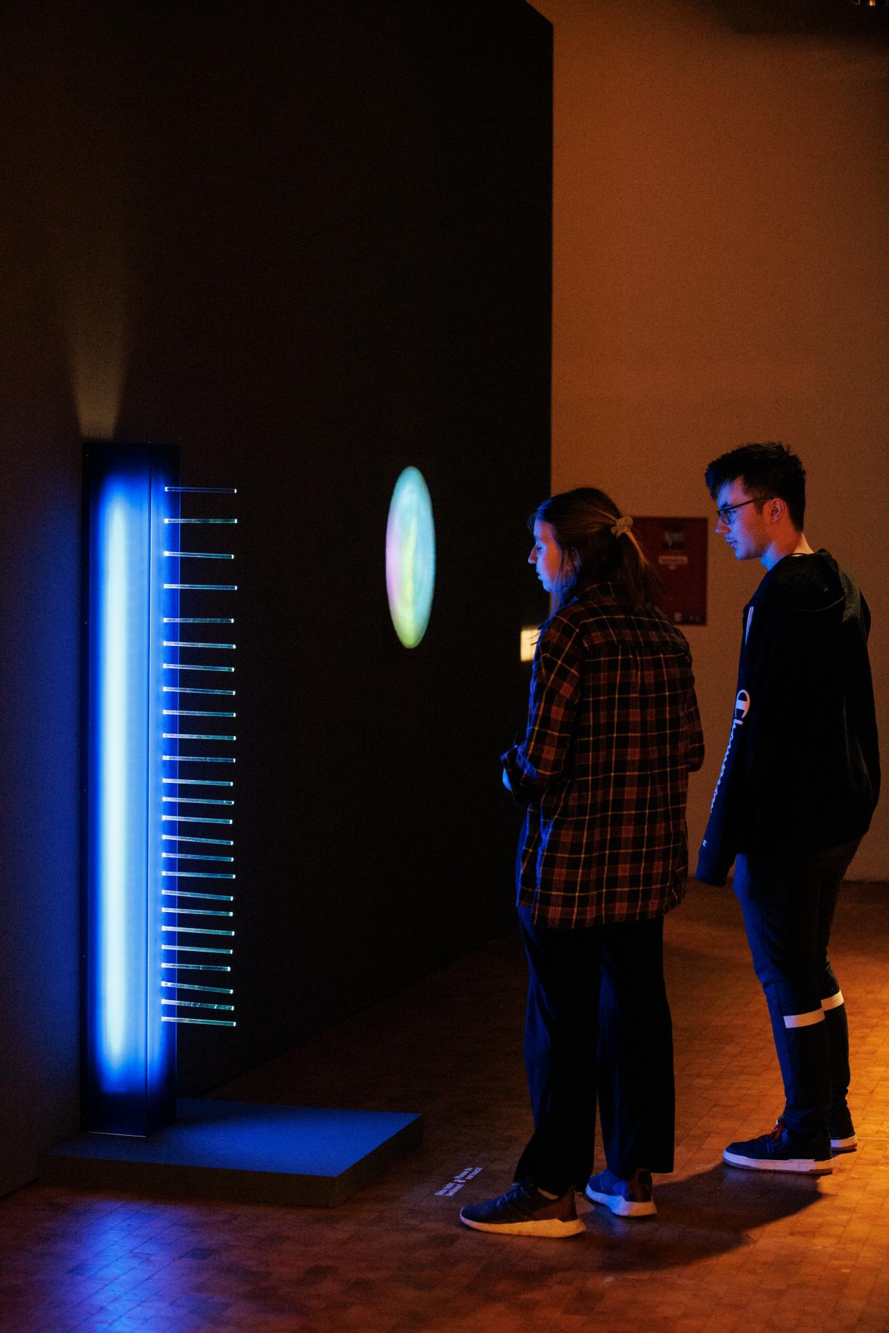 Zwei junge Menschen stehen vor einer blau leuchtenden Stele, die in etwa die Größe eines Menschen hat. Im Hintergrund leuchten verschiedenen Kunstwerke in bunten Farben auf.