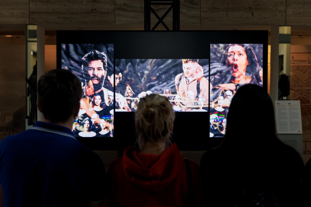 Auf dem Bild ist die Videoinstallation von John Sanborn im Foyer des rathauses zu sehen. Aud 3 Bildschirmen sind mehrere Menschen abgebildet, die unterschiedliche Expressionen ausdrücken.
