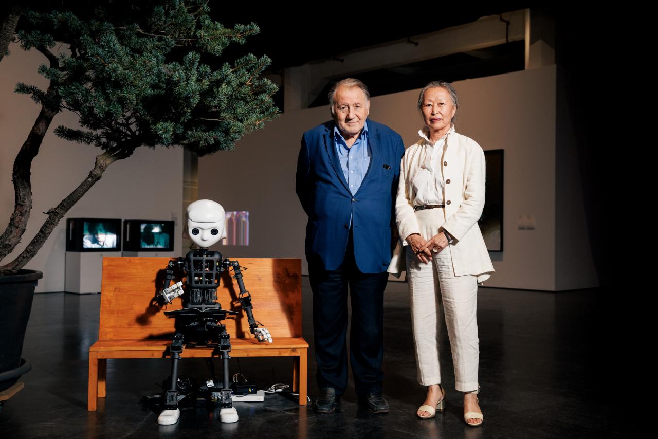 Zu sehen ist ein Roboter, dessen Aufbau dem Korpus des Menschen gleicht. Er sitzt auf einer Bank. Recht daneben steht Peter Weibel zusammen mit Soun-Gui Kim.