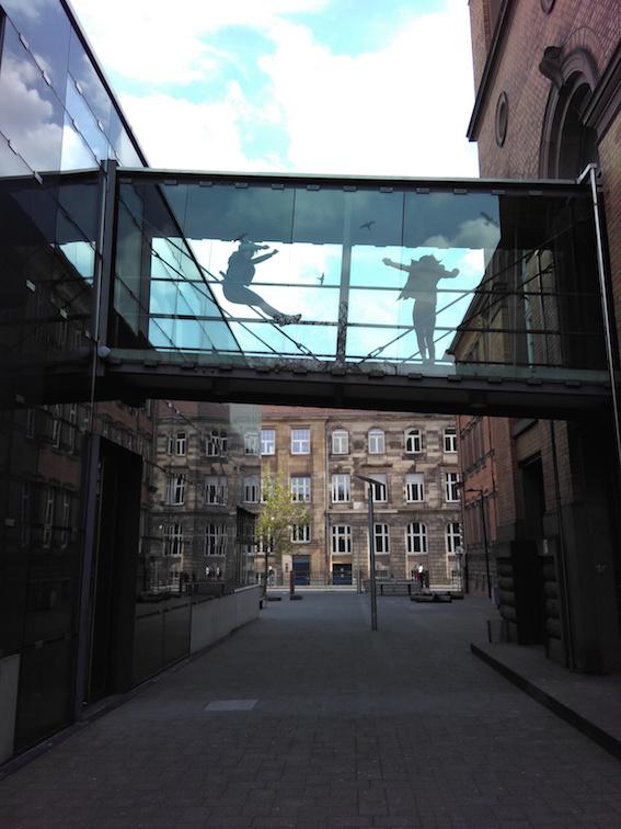 The picture shows the view into an alley with sandstone houses. Between the houses you can see a diagonally running bridge, which was covered with glass. On the bridge two persons jump, which one sees through the glass sillhouette-like.