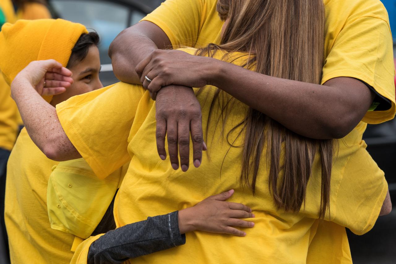Eine Frau in einem gelben T-Shirt umarmt ein Kind, welches ebenfalls gelb trägt, die Gesichter sind nicht zu sehen. Im Fokus stehen die Arme, die sich umschlingen.