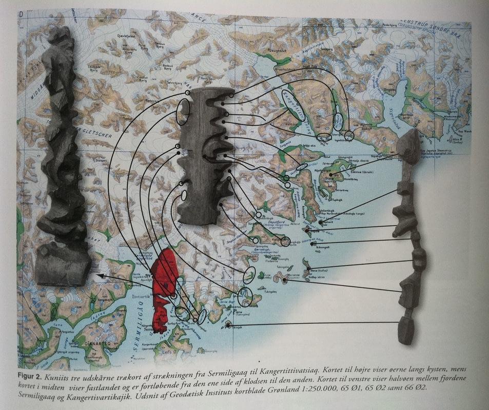 The photo shows the cartography of Greenland. On it lie various elongated wooden sculptures