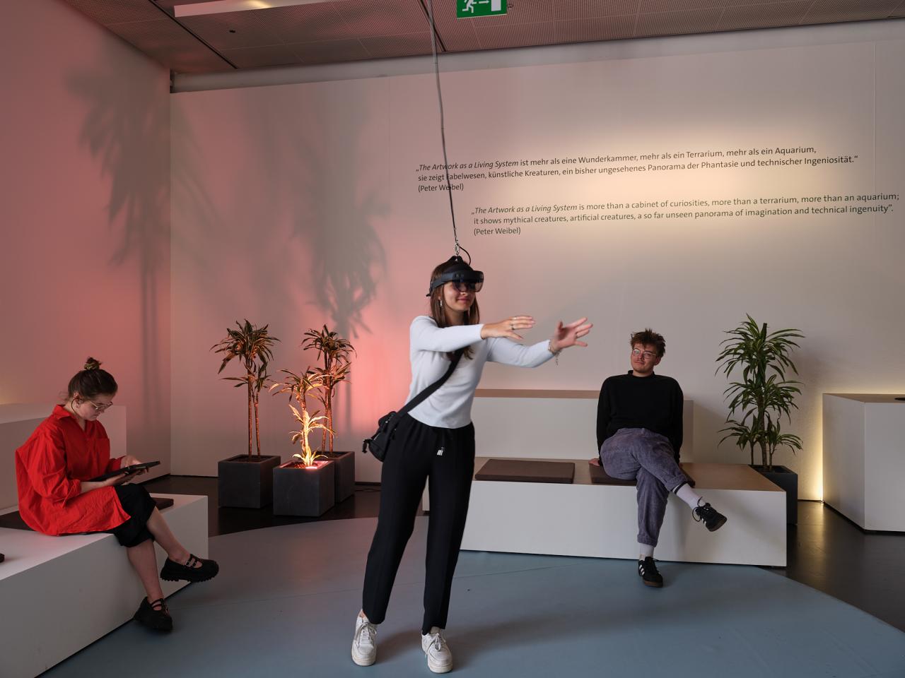On display is the work "ARtchive". The picture shows three people, two sitting on the sofas and one person standing in the middle with VR glasses on and interacting with something that only she can see.
