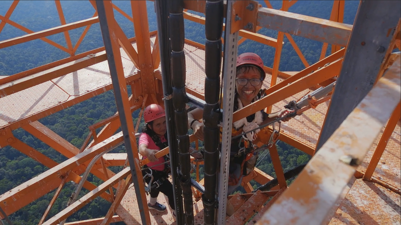 You can see two women with helmets on their heads, grinning as they climb up a steel scaffolding.