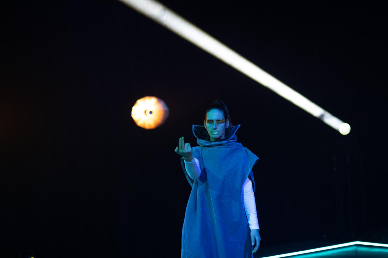 You can see a person in a blue robe against a black background. A white beam of light why a round cone of light can be seen behind her.