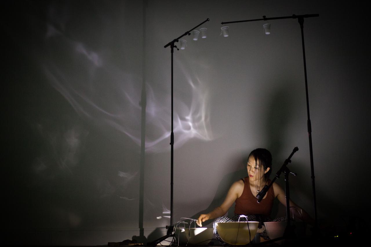 Tomoko Sauvage sitzt auf dem Boden, vor ihr stehen mit Wasser gefüllte Schalen. An der Wand sind Lichtreflexionen des Wassers zu sehen.