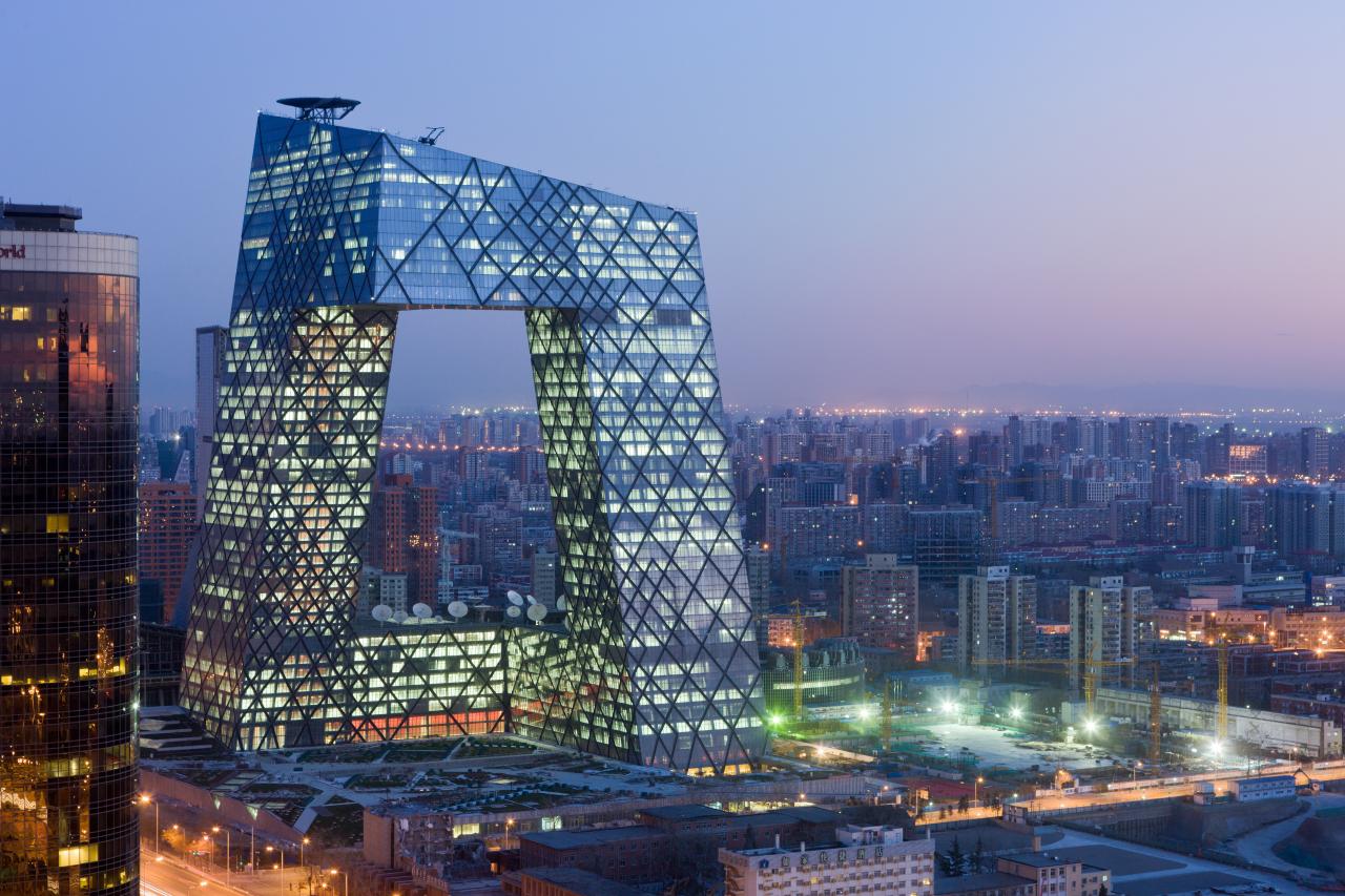 The picture shows a cityscape, at dusk. The center is a large illuminated building in the form of a gate or arch. 