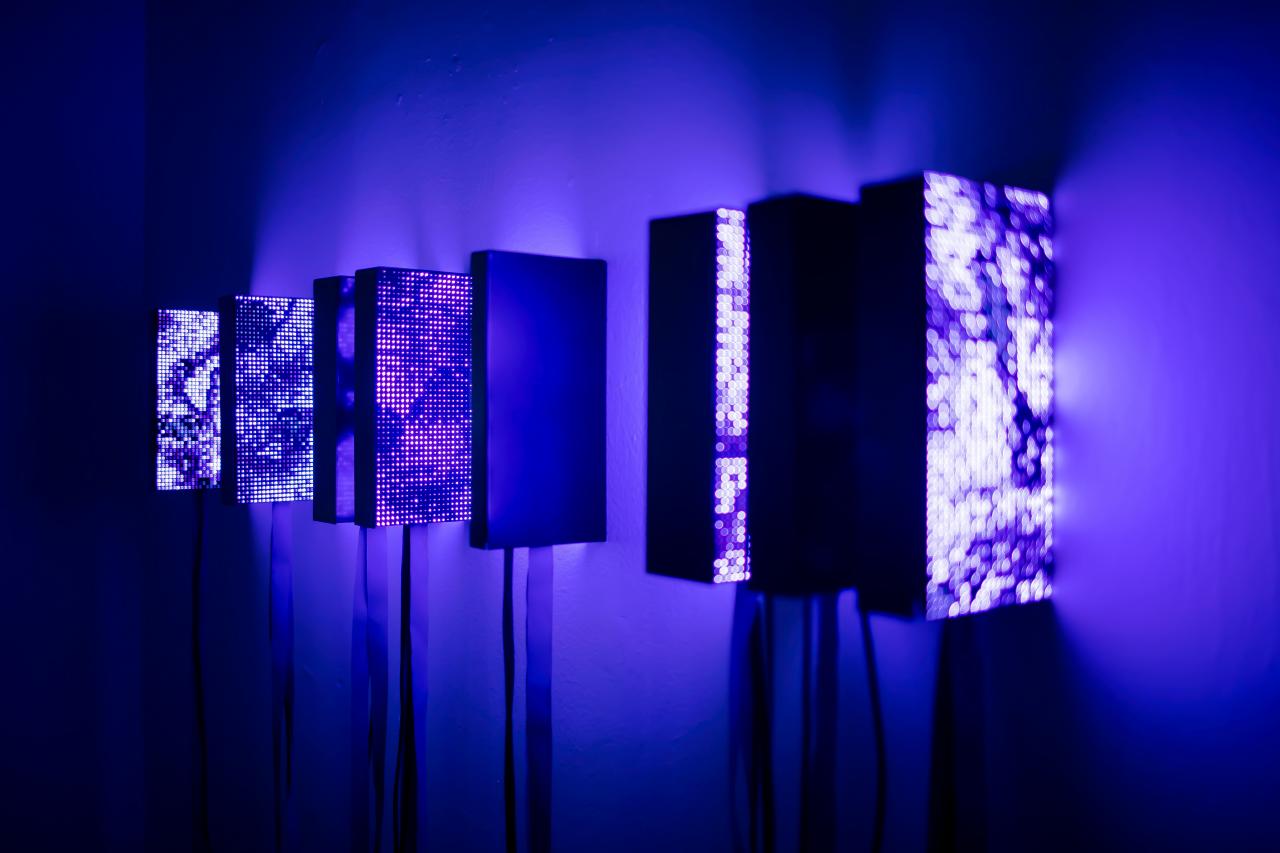 Die Installation besteht aus acht an der Wand hängenden Kästen, die zu leuchten scheinen und den Raum in eine dunkelblaue Farbe tunken.