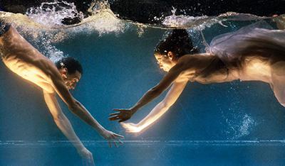Ausschnitt aus Sasha Waltz "Dido & Aeneas". Zwei halbnackte Tänzer zeigen eine Choreografie in einem bis zum Bildrand gefüllten Wassertank, welcher von unten beleuchtet wird.