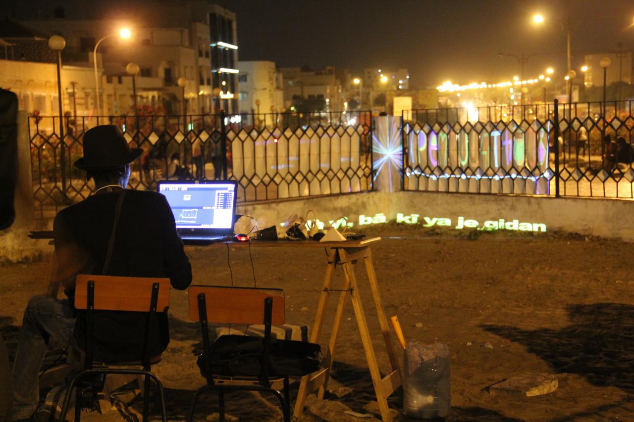 Ein Mann sitzt mit dem Rücken zu uns an einem Laptop an einenm Schreibtich auf einer von Straßenlichtern erleuchteten nächtlichen Terasse. 