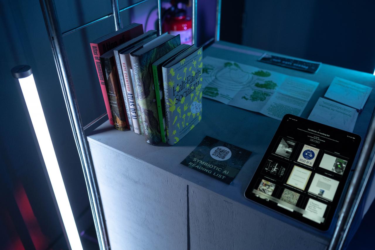 Hier sieht man das Werk »DO AIS DREAM OF CLIMATE CHAOS - SYMBIOTIC AI«. Zu sehen sind verschiedene Bücher, Flyer und ein Tablet auf einem Pult, das von der Seite beleuchtet wird.