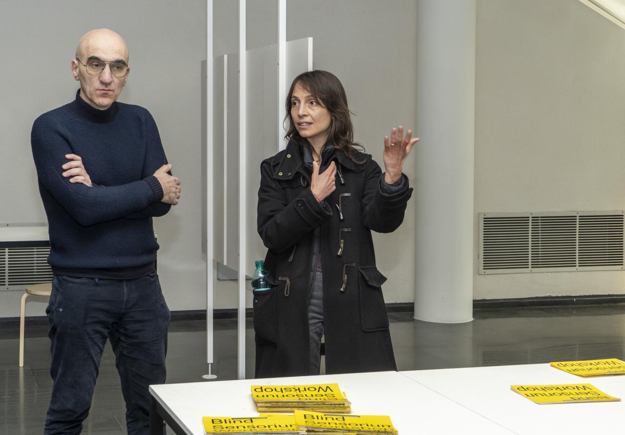 Two people stand in front of a table with yellow and black cards.