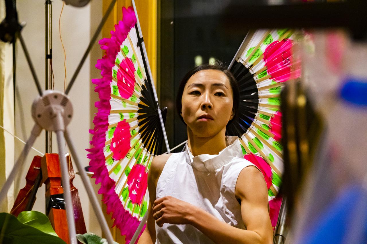 Das Foto zeigt den Oberköper und Kopf einer koreanischen Performerin die zwei pink/hellgrün farbige Fächer aus Holz als Flügel trägt.