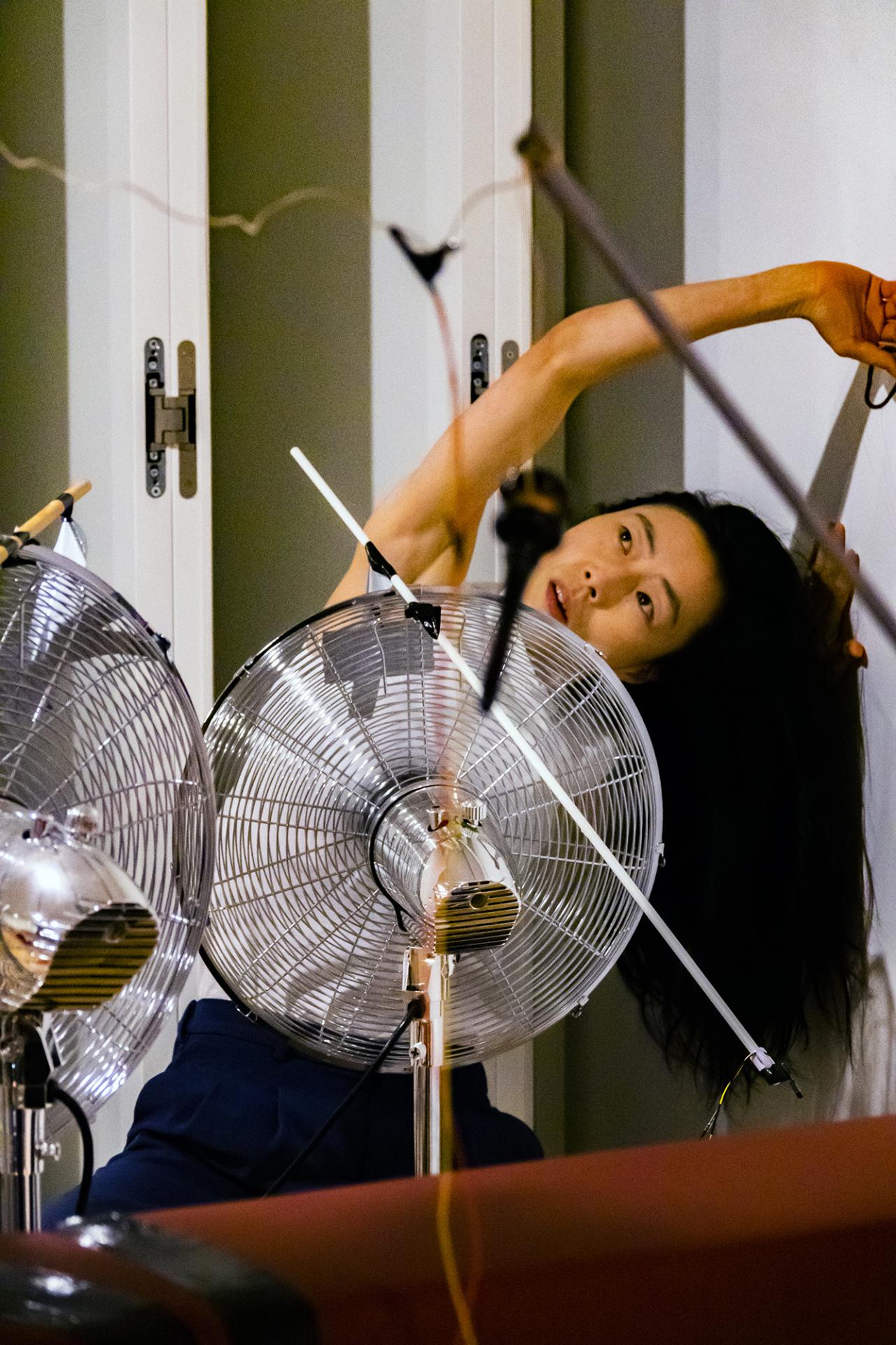 The photo shows a Korean performer bent to the left stretching her arm over her head while a large silver fan is standing in front of her.