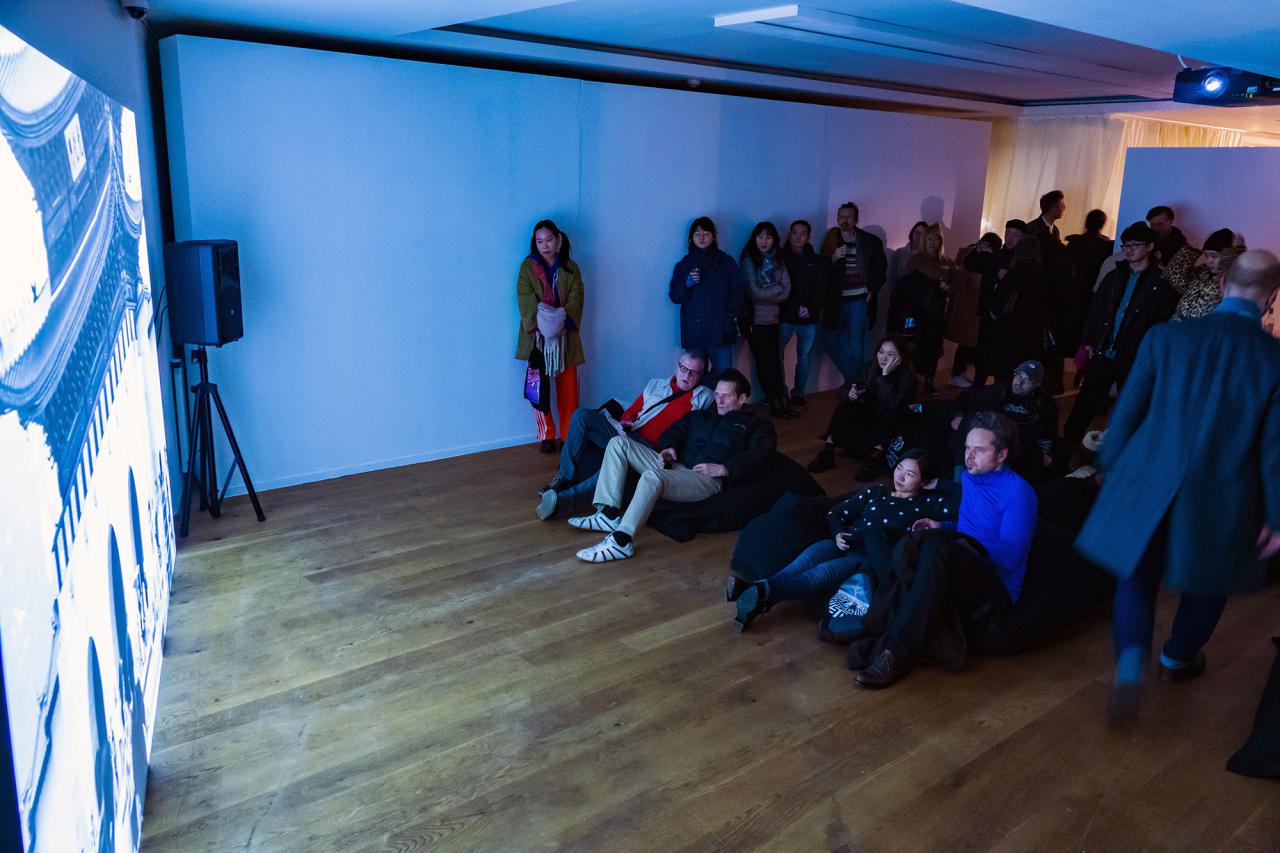 Zu sehen sind viele Menschen die vor einer großen Kunstprojektion stehen oder liegen. Der Raum hat einen Holzboden und wird von der Projektion blau erleuchtet.