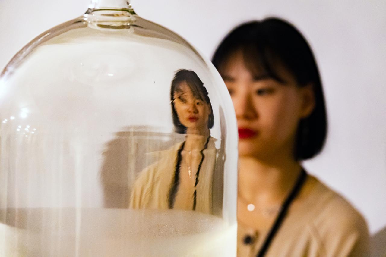 Das Foto zeigt eine Glasglocke hinter der eine koreanische Frau steht. Ihr Gesicht und Oberkörper reflektieren verzerrt in der Glasglocke.