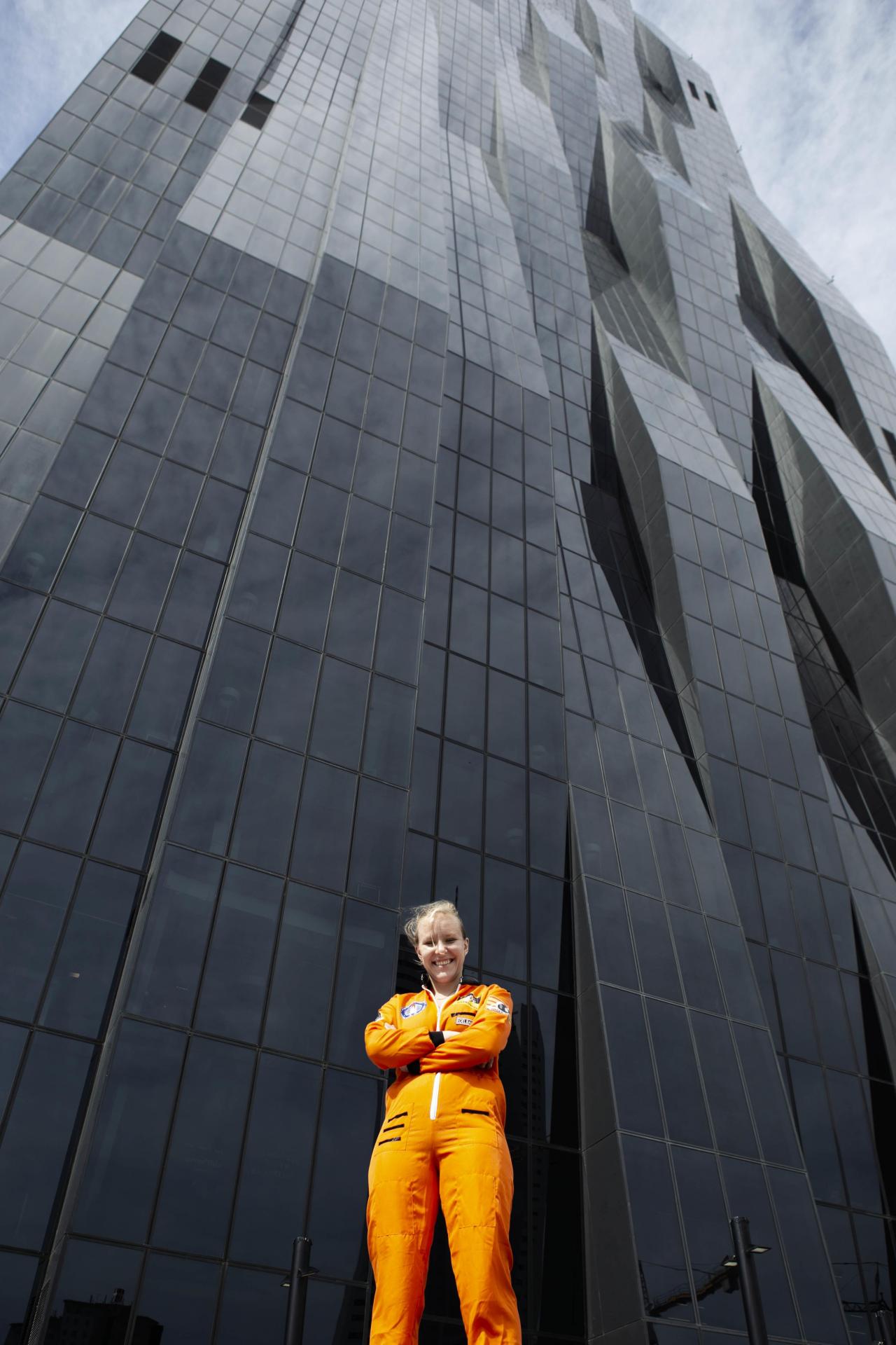 Raphaela Edelbauer in an orange space suit in front of a glass skyscraper.