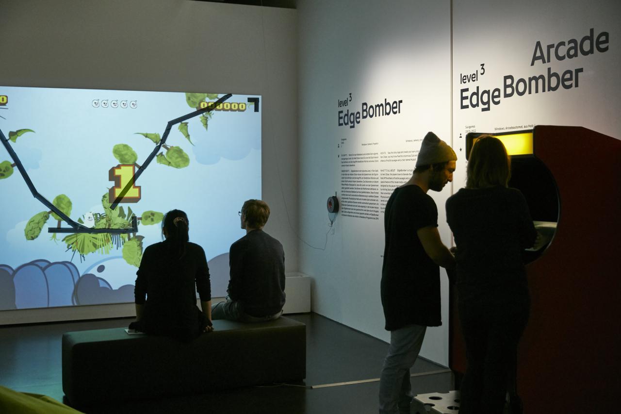 Exhibition view »EdgeBomber« with players in front of a wall projection and a game machine