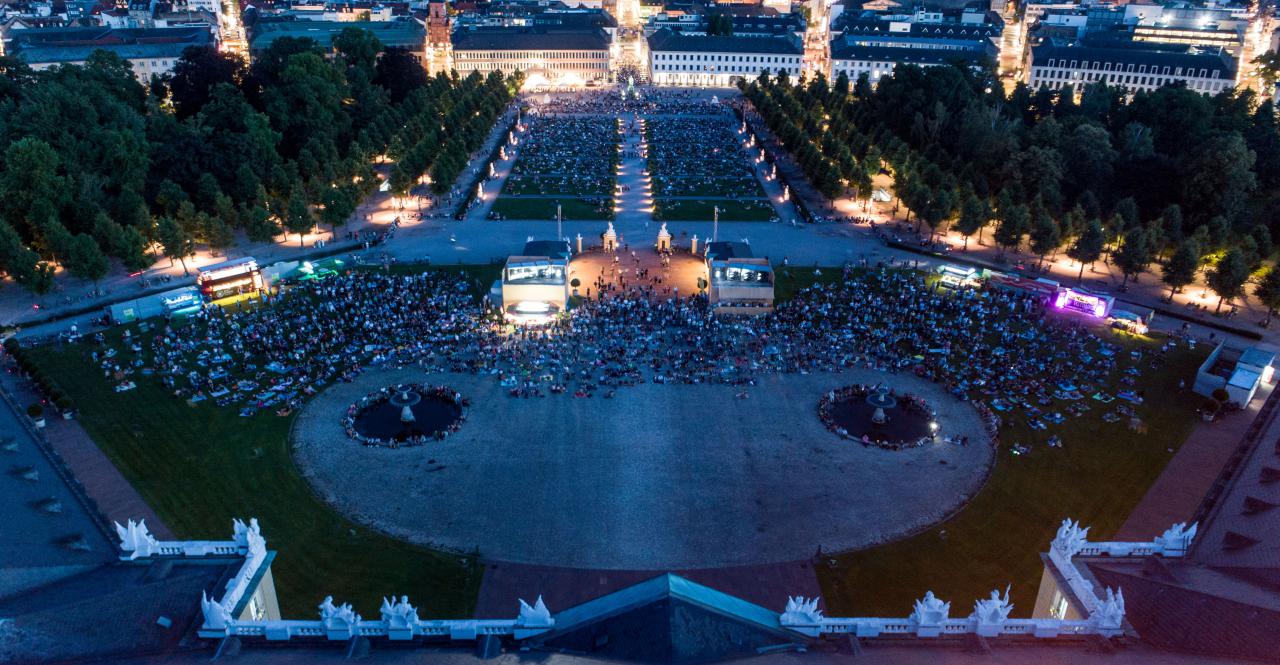 The photo shows an overview of the visitors of the premiere of the Schlosslichtspiele 2019. The photo was taken with a drone above the castle and shows a large crowd of people on the green meadow in front of Karlsruhe Castle.