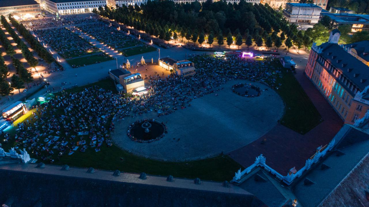 The photo shows an overview of the visitors to the premiere of the Schlosslichtspiele 2019. The photo was taken with a drone above the castle and shows a large crowd of people on the green meadow in front of Karlsruhe Castle.
