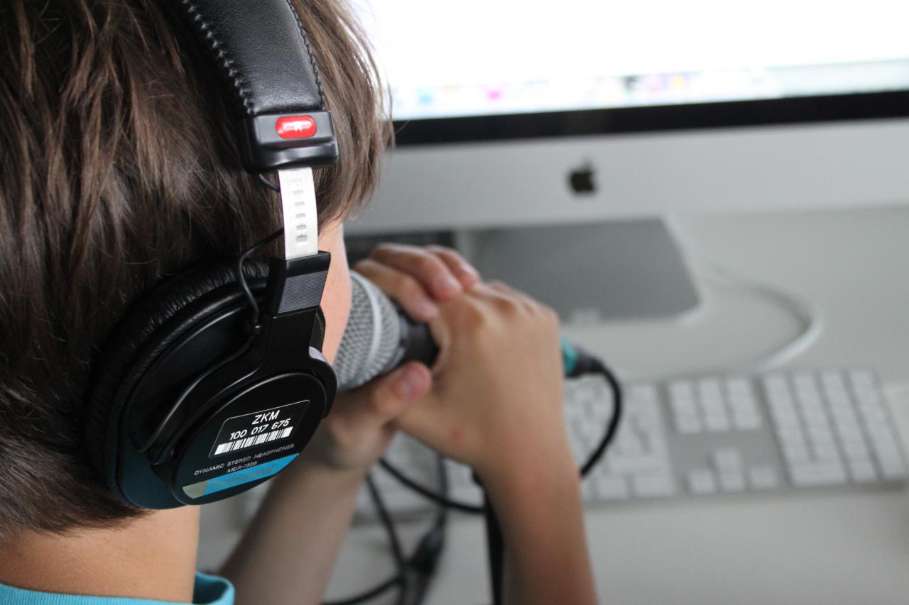 Ein Junge spricht in ein Mikrofon, das er mit beiden Händen vor den Mund hält. Er schaut dabei auf den Computerbildschirm vor ihm.