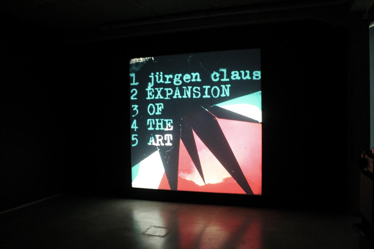 Foto einer Multimediaprojektion, ein Bild in Rosa, Schwarz und Türkis mit geometrischen Formen. Darauf der Text: »Jürgen Claus, Expansion of the Art«