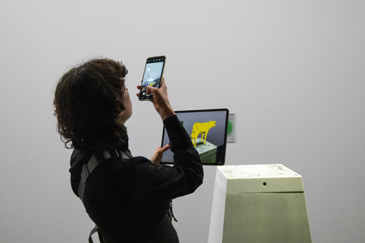 Ausstellungsansicht »Matter. Non-Matter. Anti-Matter« im ZKM | Zentrum für Kunst und Medien Karlsruhe, 2022. Eine Person fotografiert mit ihrem Handy ein Tablet, auf dem eine gelbe Kuh zu sehen ist.