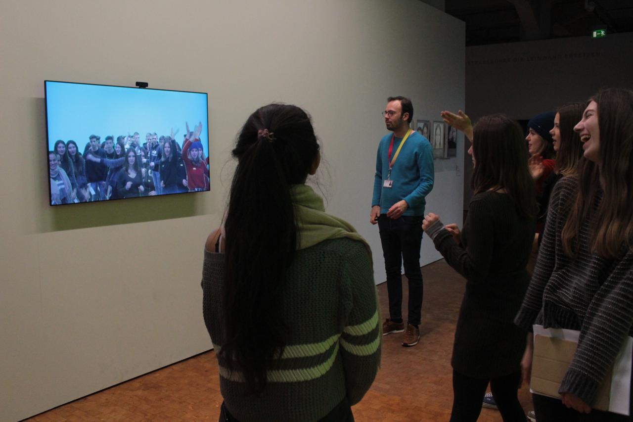 Zu sehen ist eine Gruppe an jungen Menschen, die vor einem Bildschirm stehen. Auf dem Bildschirm ist ein Live-Bild von der Gruppe zu sehen, das von einer Kamera über dem Bildschirm aufgenommen wird.