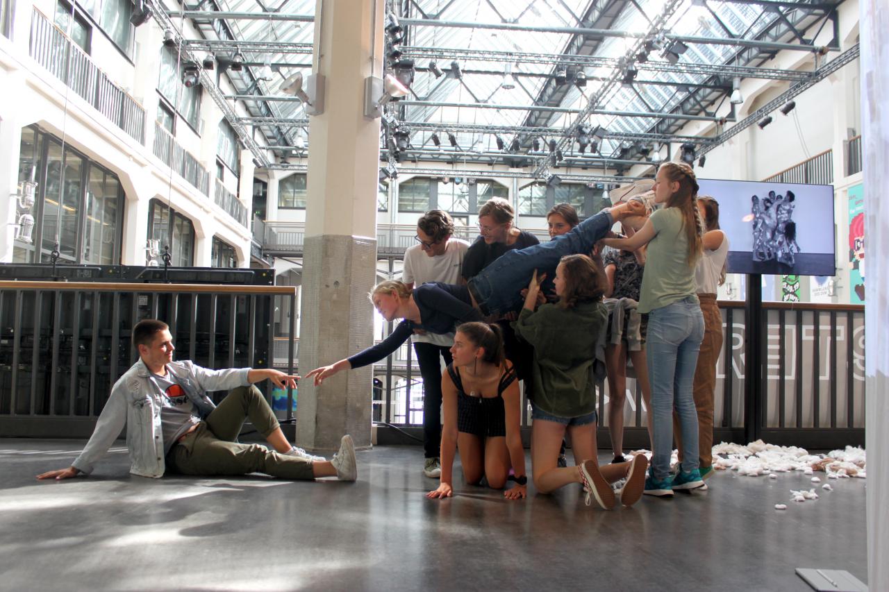 Rechts hält eine Gruppe von Schülern eine Schülerin, die ihre Hand zu einem sitzenden Schüler links streckt im Rahmen einer Veranstaltung der Kulturakademie.