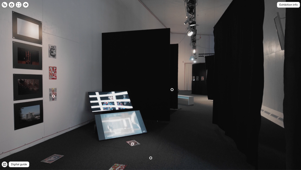 View into an exhibition room, which is hung with room-dividing black curtains. On the left side of one wall hang small photographs of paintings and colorful drawings with large letters. Two screens in the room show various videos, apparently of performanc