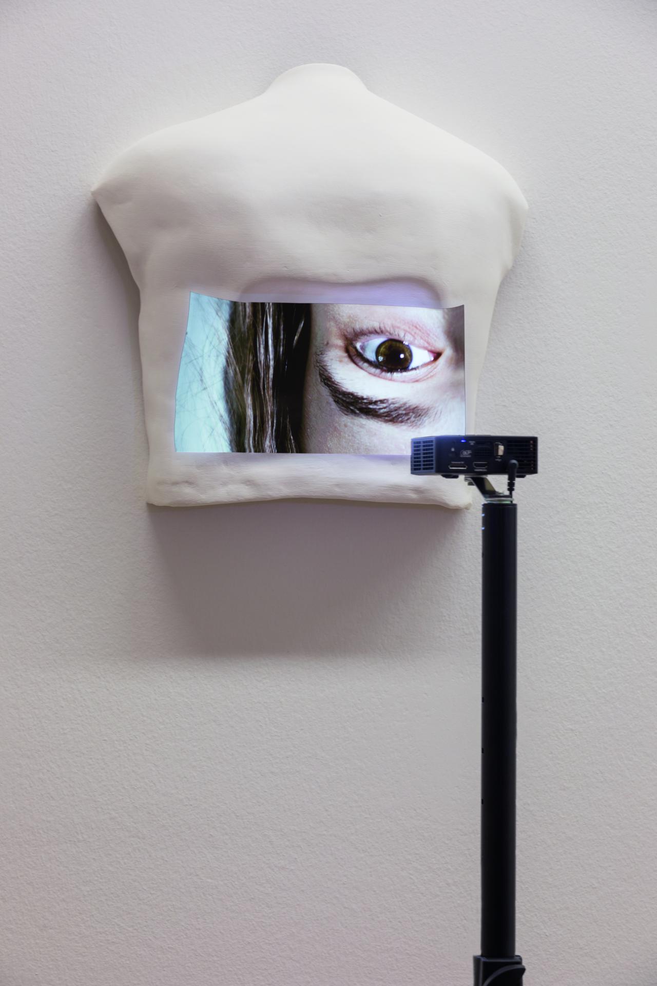 Zu sehen ist ein weißes Objekt, welches an der Wand hängt. Auf dem Objekt ist das Bild eines Abschnitts aus dem Gesicht einer Frau. 