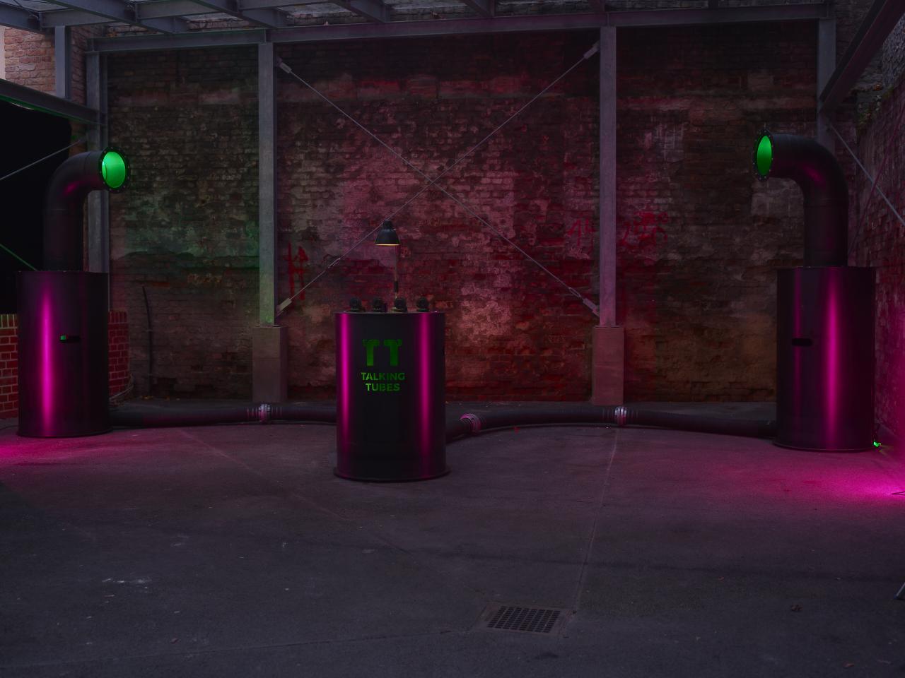 A red-lit room with the Talking Tubes installation: a control panel between two large tubes
