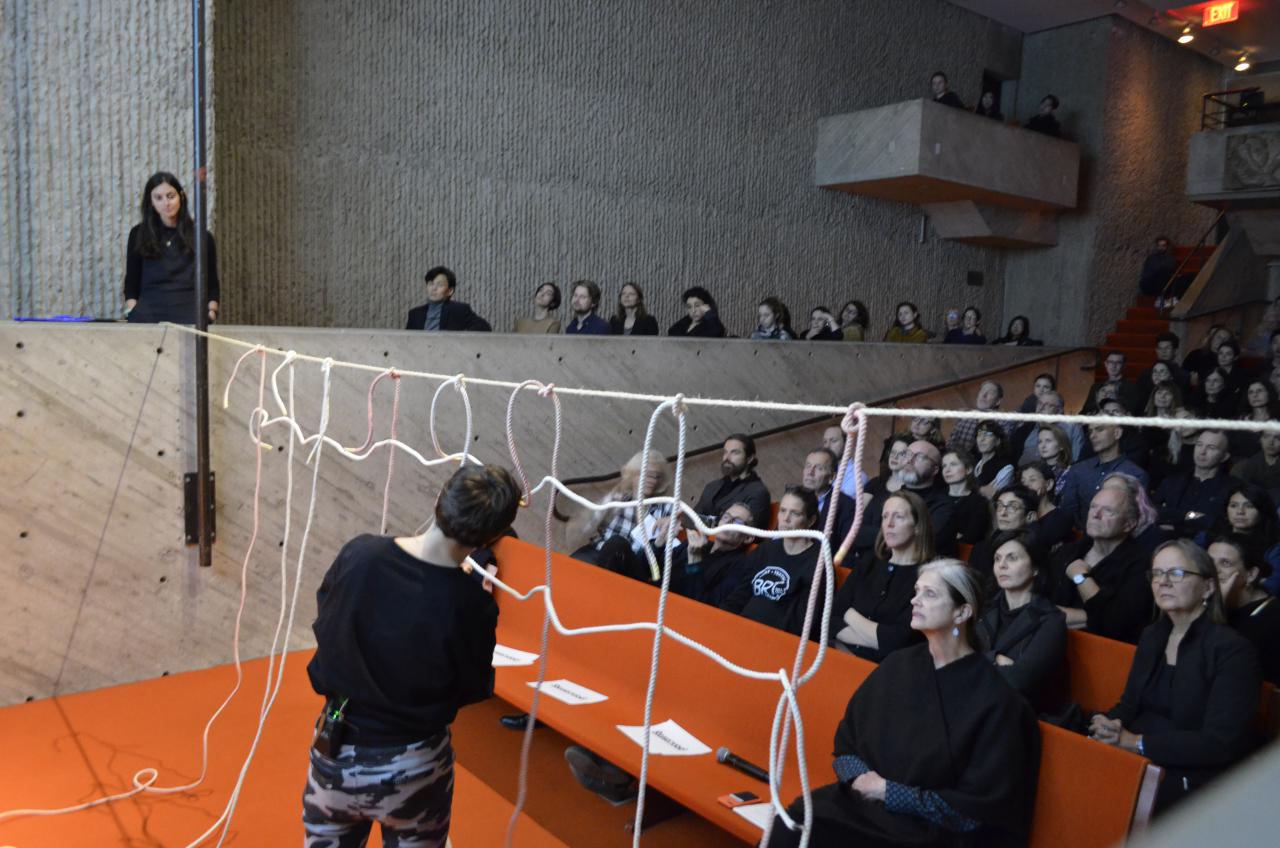 The photo shows a brutalist lecture hall with a view of the audience. The stage is cut. A large audience watches the performer form a net of ropes.