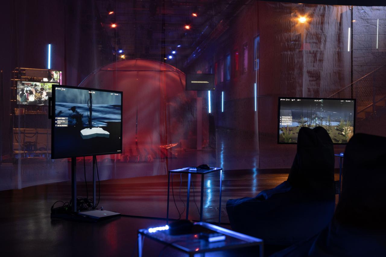 Here you can see the work »Monsters and Ghosts of the Far North«. You can see two screens on which video games are running. In front of them you can see tables with controllers to control the video games. On the right side of the photo there are seats.