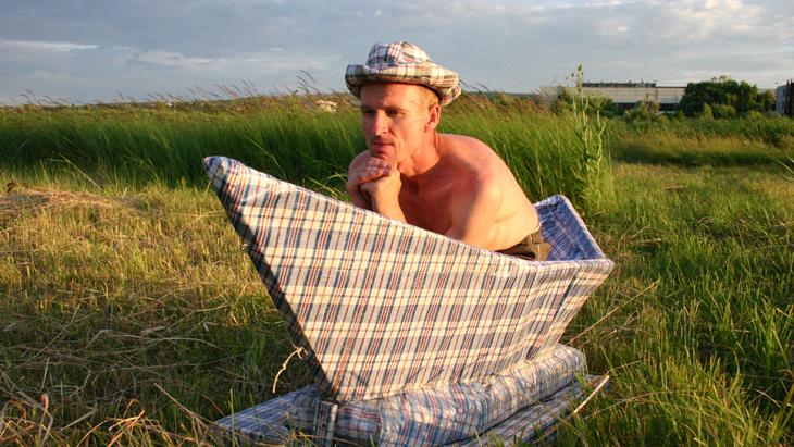 A man who is naked from the waste up sits in an oversized paper boat which stands on a green