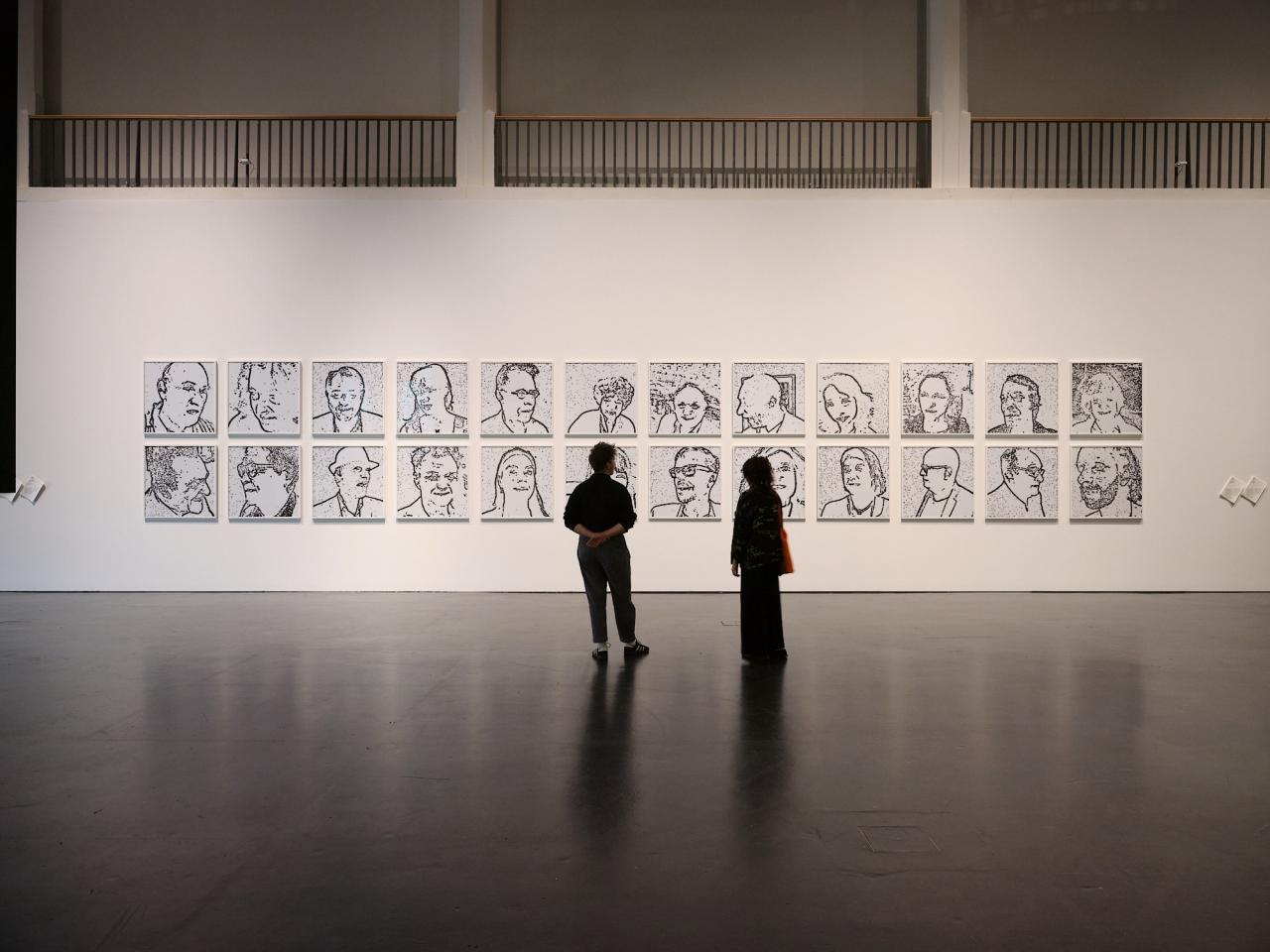 Das Bild zeigt eine Totalaufnahme des Werkes »Porträt on the Fly«, vor dem zwei Personen mit dem Rücken zur Kamera stehen und die Bilder betrachten. Im gesamten sind zwei Reihen mit je 12 Bildern zu sehen, die alle das Porträt einer Person zeigen.